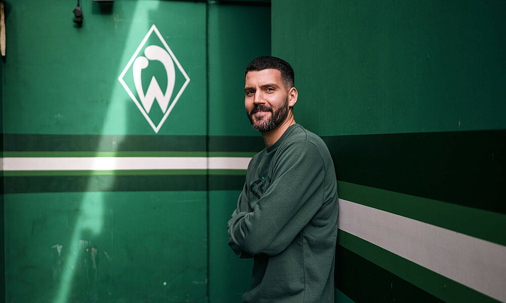 Anthony Jung leaning against a wall and smiling. In the background is the Werder logo.
