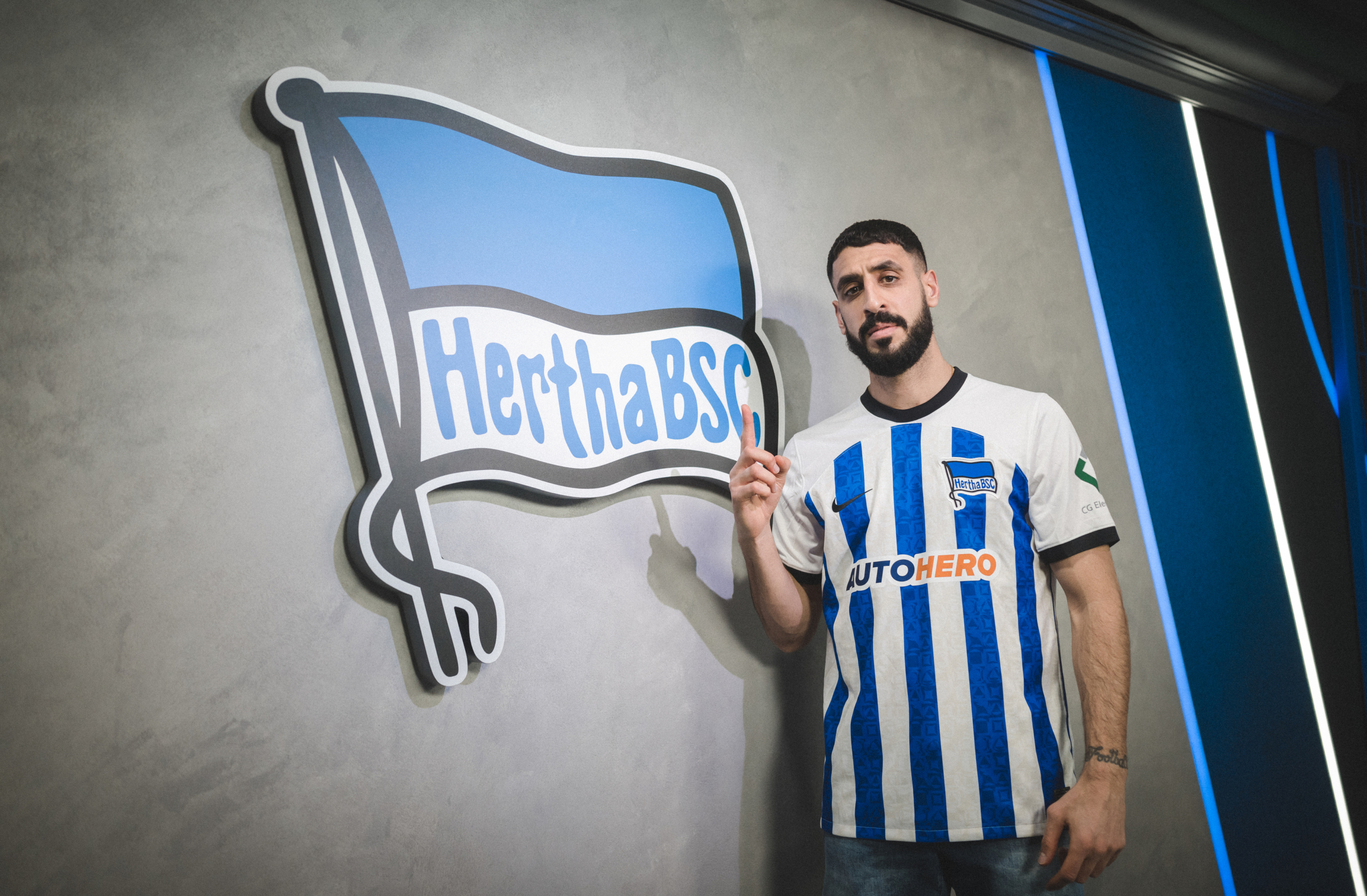 Tolga Ciğerci poses with a Hertha shirt in front of the flag.