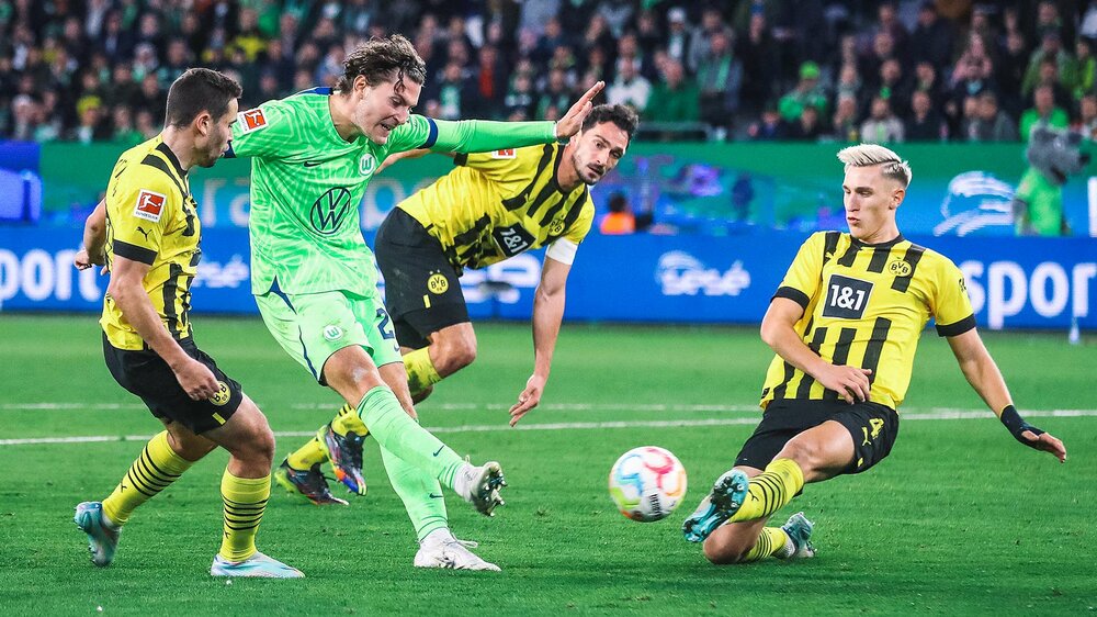 VfL-Wolfsburg-Spieler Jonas Wind setzt sich gegen Gegenspieler durch und schießt den Ball.