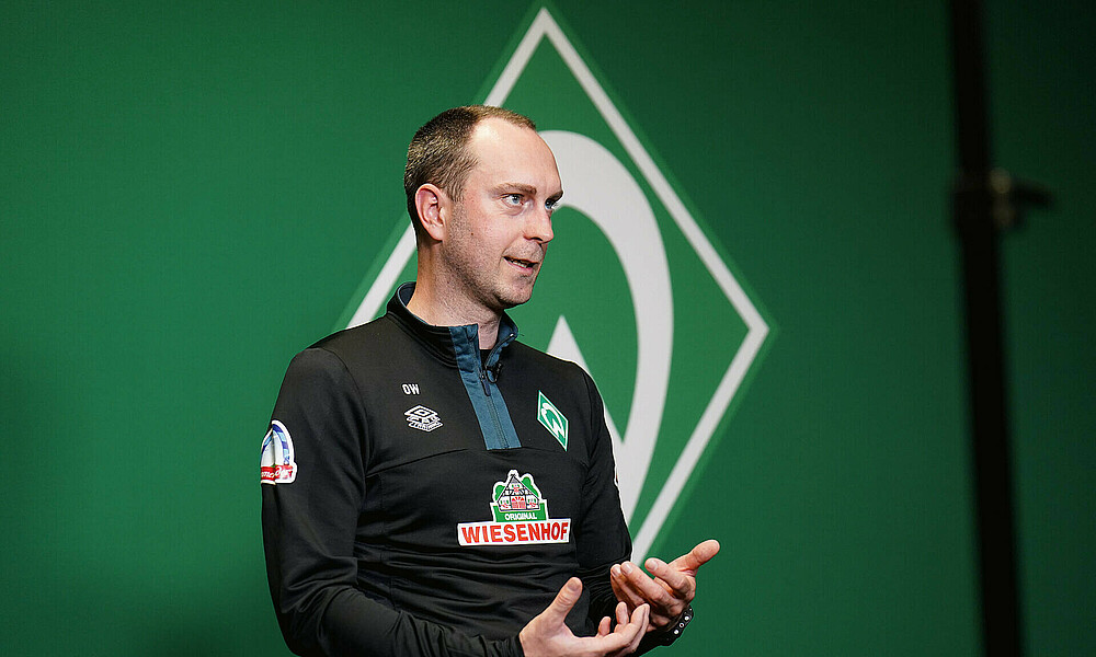 Ole Werner sitting on a chair with the Werder crest in the background.