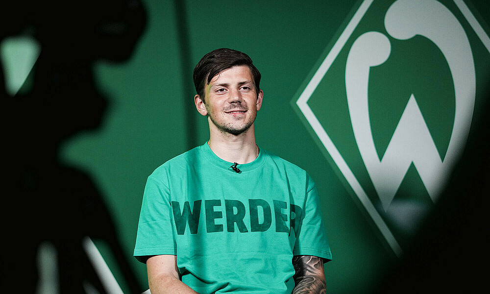 Dawid Kownacki sitting giving the interview, with a camera on the left in the foreground and the Werder crest in the background.