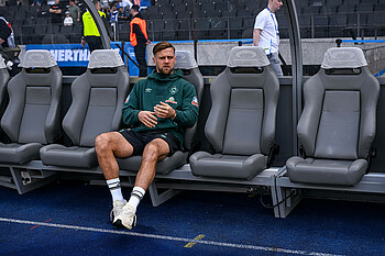 Niclas Füllkrug sitting alone on the substitutes bench at the Olympiastadion in Berlin. He is wearing a green hoodie.