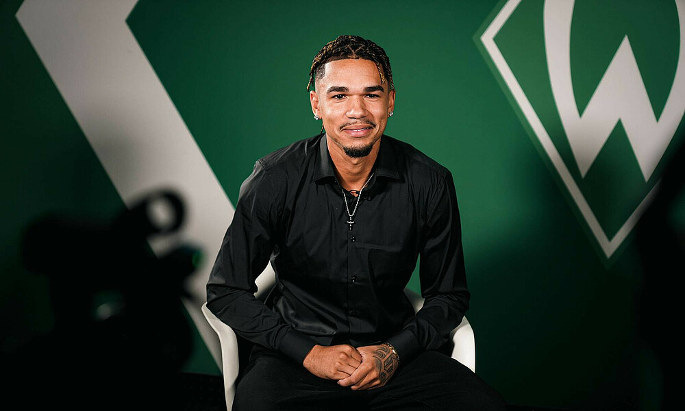 Justin Njinmah during his interview, sitting on a white chair in front of a green wall with the Werder crest on it.