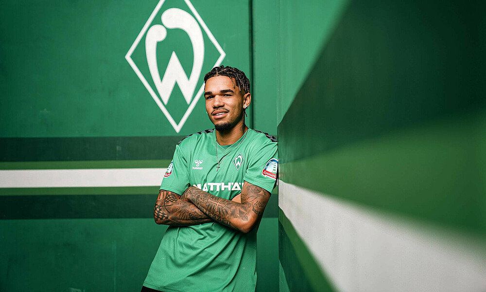 Justian Njinmah wearing a green t-shirt and leaning against a green wall with folded arms. The Werder crest is in the background.