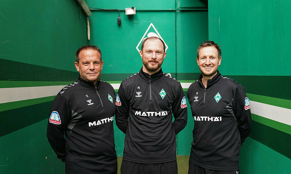 The three assistant coaches in front of a green wall.