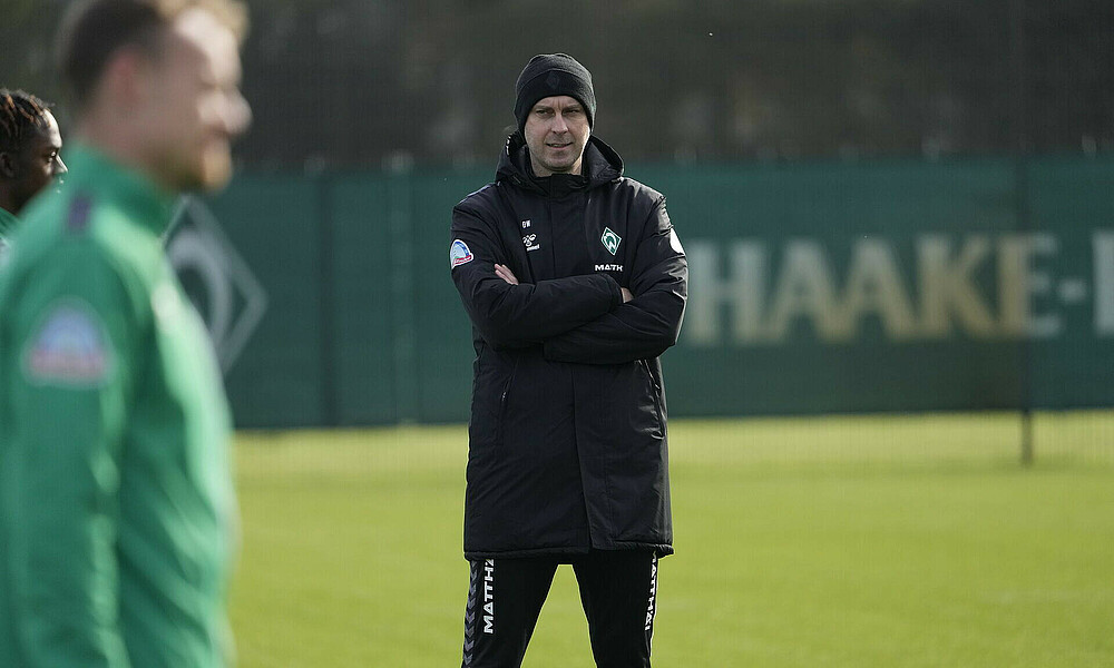 Ole Werner stands on the training pitch with his arms crossed. He is wearing a black hat and a black jacket.