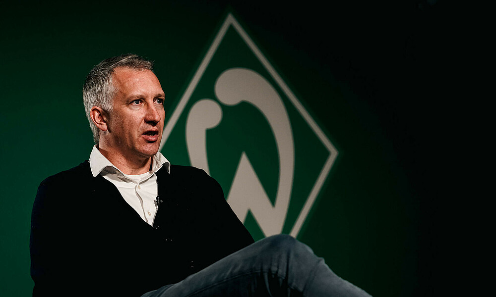 Frank Baumann sits on a chair with a large Werder crest in the background.