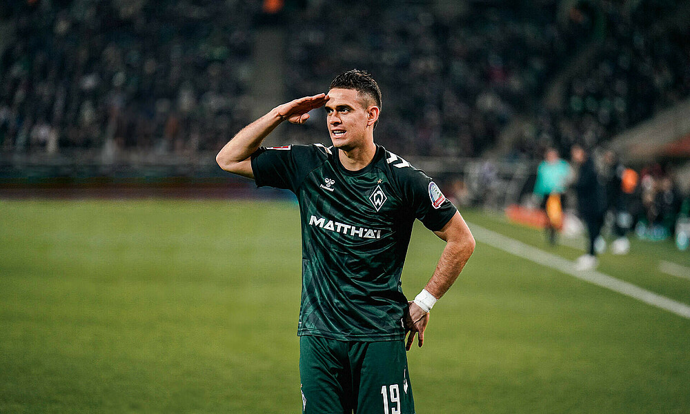 Rafael Borre salutes with his right hand with his left hand tucked behind his back. He's wearing the Werder third shirt.