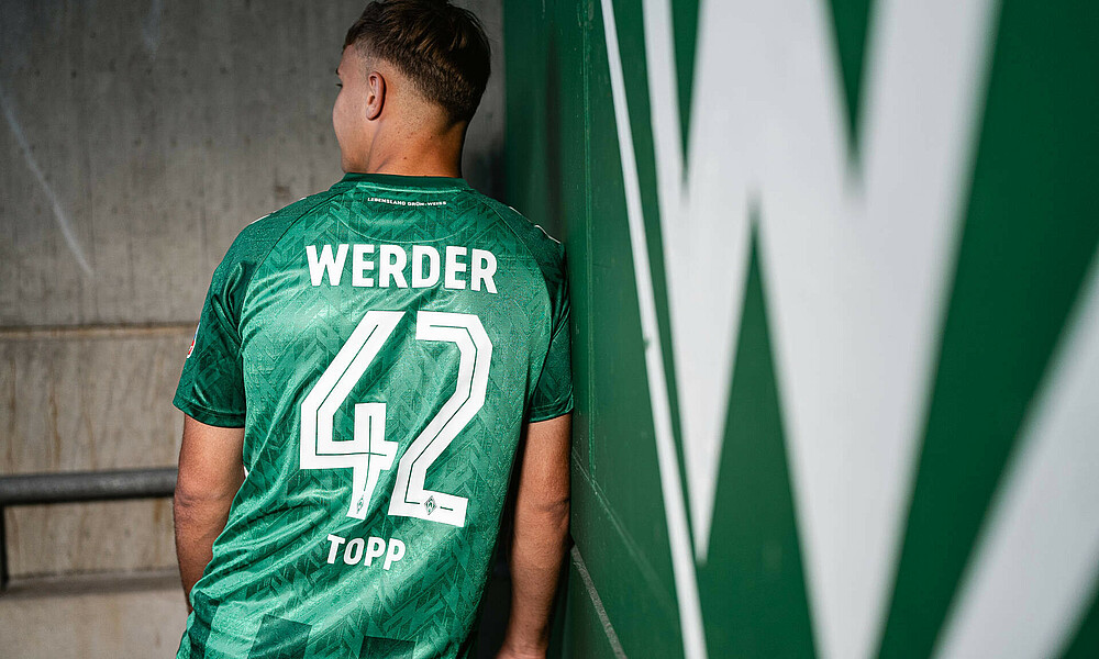 Keke Topp wearing the new Werder home shirt. He's leaning against a wall and showing his back to the camera. The back of the shirt features the number 42.