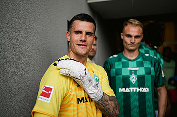 Michael Zetterer leans against a wall. He is wearing the yellow goalkeeper shirt. He is also wearing goalkeeper gloves, and one of his hands is touching his shoulder.