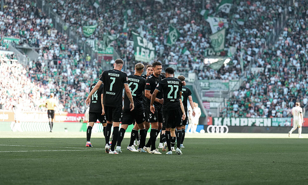 The players stand together in a group, wearing the black away kit. In the background, the Werder fans are visible. There is lots of colour.